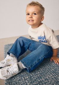 Tricou bej din bumbac cu imprimeu albastru "High Five", blugi din denim albastru, adidași albi New Balance din plasă, stând pe un covor texturat albastru.