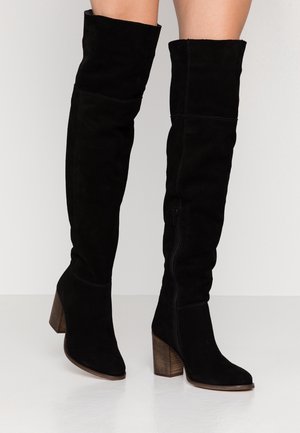 Bottes hautes en daim noir avec talons blocs en bois portées sur jambes nues sur un fond clair uni.