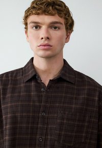 Braun kariertes Hemd mit Knopfverschluss und Kragen, ausgestattet mit schwarzen Knöpfen und einer Brusttasche. Weicher Stoff mit einem Schachbrettmuster.