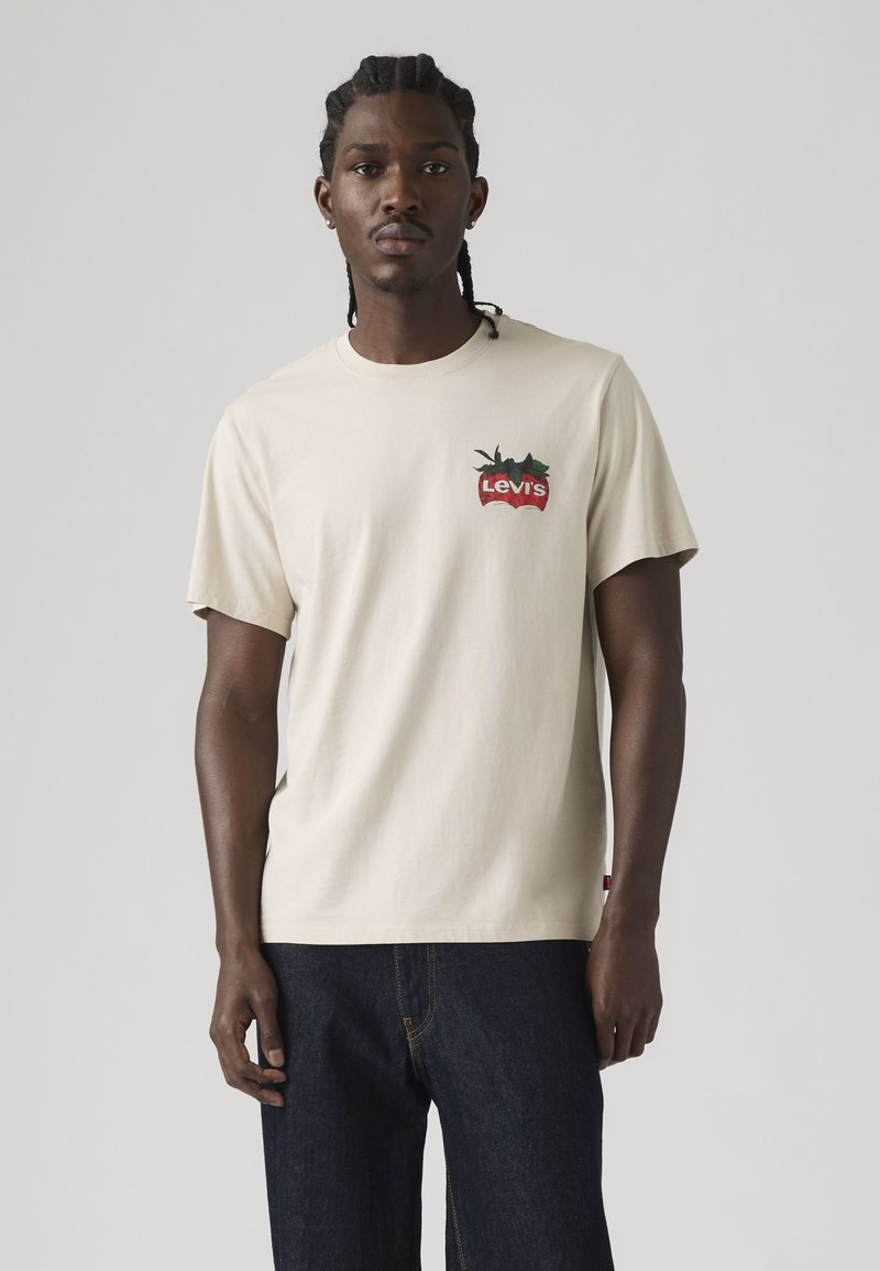 Hombre con el cabello trenzado, vistiendo una camiseta beige de Levi's con logo rojo y vaqueros azul oscuro, de pie frente a un fondo gris liso.
