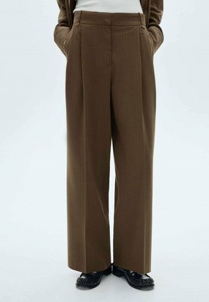 Bruine wijde broek met hoge taille en krijtstrepen, gedragen met zwarte schoenen en een wit shirt in de broek gestopt, handen in de zakken.