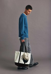 Suéter azul, camiseta blanca, jeans grises. Bolsa de lona con detalles negros y gorra. Pose de pie, pie izquierdo hacia adelante. Fondo neutro.