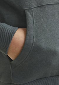 Temno siva majica s kapuco z ribastimi manšetami in sprednjim žepom. Teksturovana tkanina; prikazuje roko, ki počiva v žepu, s čimer poudarja udobje in priložnostni slog.