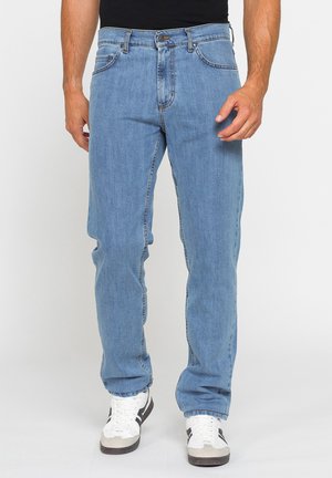 Uomo che indossa jeans azzurri a gamba dritta e sneakers bianche, in piedi davanti a uno sfondo bianco.