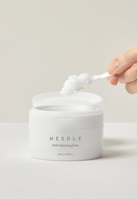 Valkoinen sylinterimäinen astia kannella, jossa lukee "NEEDLY Mild cleansing balm". Astiassa on paksu, koostumukseltaan valkoinen balsami valkoisella lastalla.