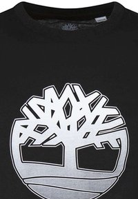 T-shirt noir avec un grand motif graphique blanc représentant un logo d'arbre stylisé. Le logo présente des lignes audacieuses et des formes géométriques.