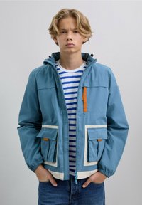 Chaqueta azul claro con detalles en blanco y cremallera naranja. Tiene dos bolsillos frontales y una capucha. Lleva puesta sobre una camisa de rayas.
