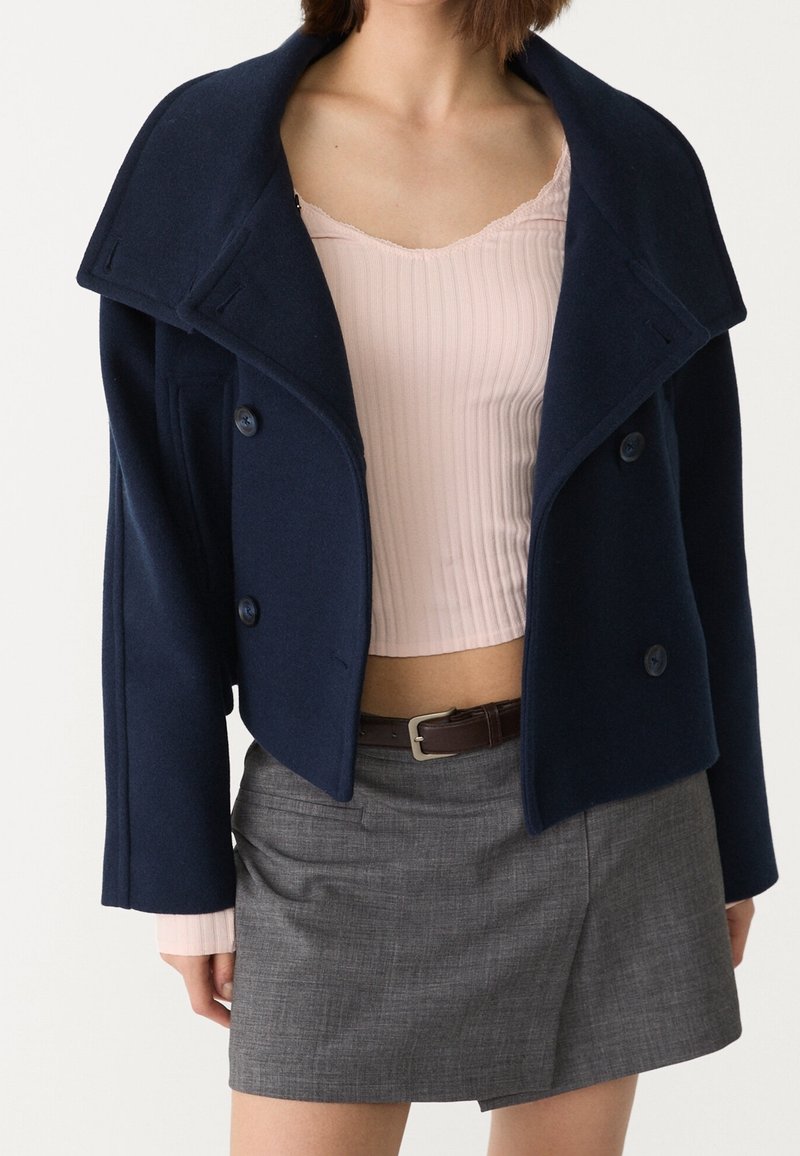 Femme portant un manteau court croisé bleu marine, un haut côtelé rose clair, une mini-jupe grise et une ceinture foncée sur un fond uni.