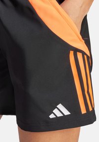 Schwarze Sportshorts mit auffälligen orangefarbenen Akzenten und einem Logo mit drei weißen Streifen. Hergestellt aus feuchtigkeitsableitendem Material mit glatter Textur.