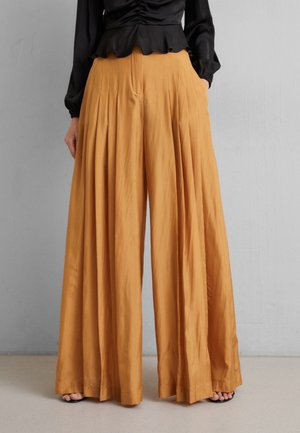 Woman wearing high-waisted, wide-leg mustard pants with pleats, paired with a black long-sleeve top, standing against a plain gray wall.