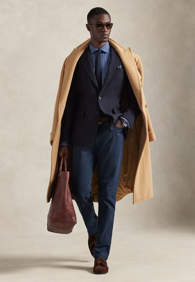 Blazer azul-marinho sobre uma camisa xadrez azul-claro, casaco caramelo, calças escuras, mala de couro castanho e mocassins de camurça castanhos. Óculos de sol realçam o visual.