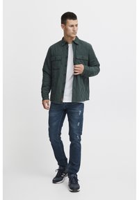 Cazadora de camisa acolchada de color verde oscuro con dos bolsillos frontales y cierre de botones, llevada sobre una camiseta blanca, combinada con jeans azul desgastados y zapatillas negras.