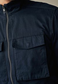 Granatowa kurtka z pionowym metalowym zamkiem błyskawicznym i dwoma dużymi kieszeniami z klapami na piersi, widoczne detale przeszyć na tkaninie.