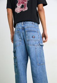 Pantalons cargo en denim bleu clair avec une coupe décontractée, plusieurs poches et une étiquette près de la taille arrière. Comprend des coutures contrastantes.