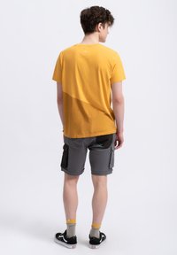 Geel t-shirt met een diagonaal kleurblokpatroon, gecombineerd met grijze cargoshorts met zijzakken en zwarte accenten.