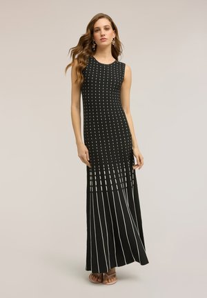 Frau steht in einem ärmellosen schwarzen Maxikleid mit vertikalen weißen gepunkteten und gestreiften Mustern, trägt hängende Ohrringe und Sandalen.