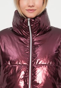 Metalická burgundy prošívaná bunda s vysokým límcem, předním zipem a prošívanou texturou. Hladký, reflexní materiál se strukturovanými designovými detaily.