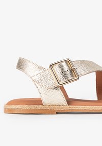 Sandalias con tiras en faux cuero dorado metálico, con una gran hebilla cuadrada, una plantilla en tono marrón, y un borde de yute trenzado a lo largo de la suela.
