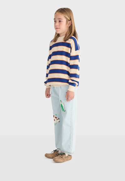 Enfant debout, portant un pull à rayures beige et bleu, un pantalon bleu clair avec des illustrations imprimées et des chaussures décontractées marron sur un fond uni.
