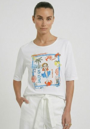 Femme aux cheveux plaqués en arrière portant une chemise blanche avec des illustrations colorées sur le thème de la plage et du texte, associée à un pantalon blanc et des bagues.