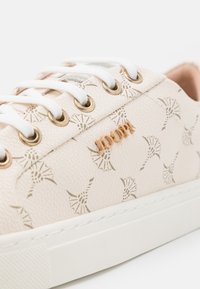 Zapatillas beige con una superficie texturizada, que presentan un patrón sutil, cordones blancos, ojales dorados y un pequeño acento de logo de bronce.