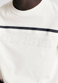 Camiseta de algodón blanca con cuello redondo, que presenta una franja azul marino y el texto en relieve "HILFIGER" en el pecho. Textura suave y corte relajado.