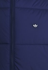 Marinblå pufferjacka med dragkedja i fronten, med horisontell sömnad och en broderad logotyp i vitt på vänster bröst. Slät yta.