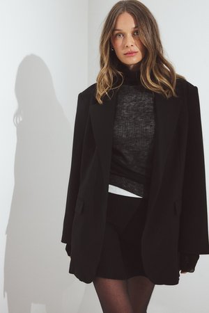 Melna lielizmēra žakete ar iegrieztām atlokām, valkāta virs teksturēta tumša turtlenecka un melnas svārku. Gluda auduma ar nelielu spīdumu.
