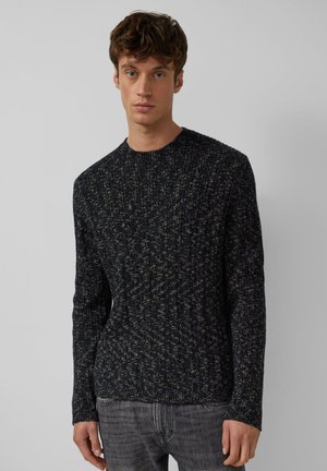 Junger Mann, der einen dunklen, gerippten Strickpullover und graue Jeans trägt und vor einem einfarbigen hellgrauen Hintergrund steht.