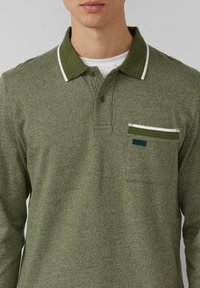 Długa koszulka polo w kolorze oliwkowej zieleni z białymi akcentami na kołnierzyku oraz kieszenią na piersi z zamkiem. Materiał o fakturze z subtelnym wzorem.