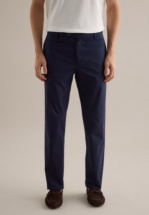 Mann trägt marineblaue Slim-Fit-Hose, weißes Hemd und braune Wildleder-Mokassins, steht vor einem einfarbigen beigen Hintergrund.