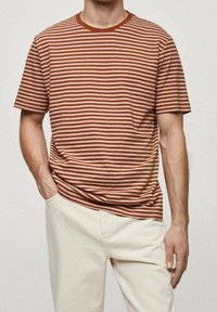 Camiseta de manga corta a rayas marrón y crema con cuello redondo, combinada con pantalones de color claro. La tela parece suave y casual.