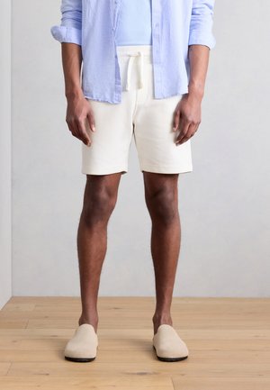 Person, die ein hellblaues Hemd mit Knöpfen, cremefarbene, strukturierte Shorts mit Kordelzug und beige Schlupfschuhe trägt, steht auf einem Holzboden.