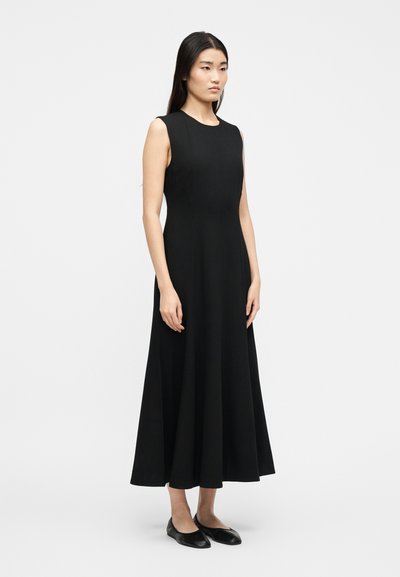 Naine seisab, kandes varrukateta musta midi kleiti ja musti tallaga kingi, taustal hele, ühtlane taust.