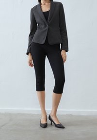 Blazer entallado gris carbón, top ajustado negro, leggings capri negros y zapatos de tacón puntiagudos sobre un fondo neutro.