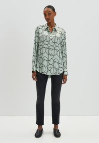 Camicia verde menta a maniche lunghe con motivi circolari neri, chiusura con bottoni e un fit rilassato, abbinata a jeans neri slim e scarpe nere.