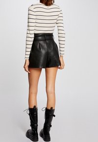 Donna che indossa pantaloni in pelle nera, stivali neri fino al polpaccio con fibbie e un top a maniche lunghe color crema con righe orizzontali nere, vista di spalle.