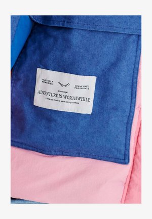 Denimová tkanina v modrých a jemně růžových vrstvách, s oblečkovou etiketou obsahující text "Dobrodružství stojí za to" a pokyny k údržbě.