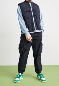 Musta kevyt liivi, jossa kaksi taskua edessä, vaaleansinisen pitkähihaisen paidan päällä, yhdistetty mustiin cargo-housuihin ja vihreisiin lenkkareihin.