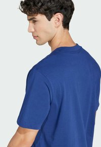 Kratke rokave modra bombažna majica s klasičnim krojem ovratnika in sproščenim fitom, ki se ponaša z gladko teksturo in čistimi šivi.