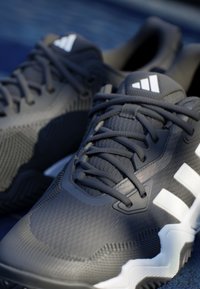 Crne sportske tenisice s teksturiranim gornjim dijelom, bijelim naglascima i čvrstim gumenim potplatom. Dizajnirane za udobnost i performanse u sportu.