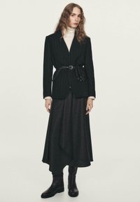 Czarna marynarka w cienkie paski ze zwężeniem w talii, noszona na kremowym golfie i asymetrycznej czarnej spódnicy, zestawiona z czarnymi kozakami za kolano.