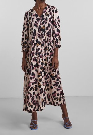 Vrouw draagt een lange beige jurk met zwarte, bruine en roze luipaardprint, driekwart mouwen en blauwe sandalen met bandjes.