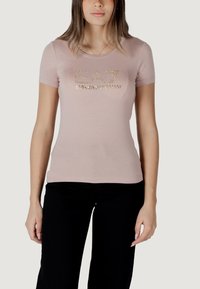 Camiseta ajustada de color rosa claro con mangas cortas y escote redondeado, que presenta un estampado de logo con tachuelas doradas en el frontal.