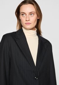 Svart pinstripet blazer med notched krage, en-knaps lukking og glatt tekstur, lagt over en beige høyhalset genser.