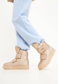 Bézs színű, vastag csizmák két széles tépőzáras pánttal, textúrált krémszínű talppal, kerek orral és sima szintetikus felsőrészsel. Mivél világos kék nadrággal viselik.