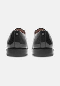 Ein Paar glänzende schwarze Leder-Oxfords von hinten gesehen, das gestickte Nähte und gestapelte Absätze auf einem neutralen Hintergrund zeigt.