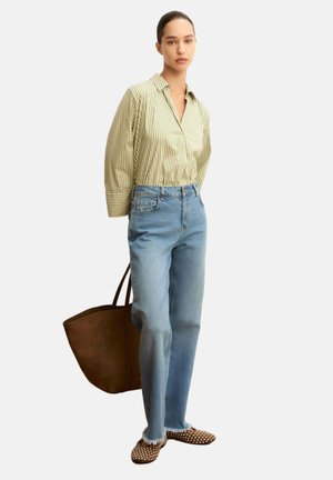 Donna che indossa una camicetta a righe gialle, jeans azzurri chiari, scarpe slip-on marroni con borchie e porta una grande borsa tote marrone.