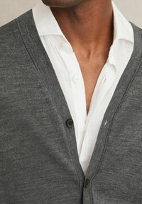 Gray knitted cardigan with a v-neck, featuring a single dark button. Worn over a white collared shirt with button details. Smooth texture.