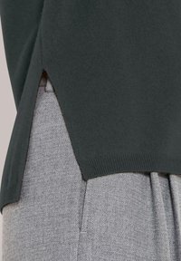 Dunkelgrüner Strickpullover mit seitlichem Schlitz über hellgrauen strukturierten Hosen mit sichtbarem Taschennaht.